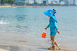© Irina Mikhailichenko - Child boy wearing blue sun protection swimwear and large blue hat walks along the beach, holding an orange bucket, city skyline background. Happy childhood concept. Summer vacation. High quality photo