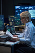 © DragonImages - Vertical shot of middle aged Caucasian woman reviewing printed report beside cybersecurity dashboards and code screens, supporting IT audit, data security, compliance marketing