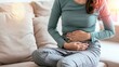 © Bonsales - Woman suffering from stomach ache, sitting on a sofa, highlighting menstrual cramps, digestion problems, or related health issues