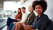 © dinatychynska - A confident young Black woman holds a smartphone and smiles at camera while diverse colleagues sit in a bright modern office, reflecting inclusive digital workplace and professional lifestyle.