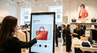 © Color Zen - A young woman interacts with a large digital touchscreen kiosk to browse clothing and handbags inside a modern bright retail boutique fashion clothing store