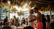 © PhotoArtHub - Two men sharing a moment under a thatched roof with palm trees and a building in the background, symbolizing a relaxed outdoor gathering or vacation scene