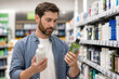 © Сергей Стельченко - A man stands in a store aisle holding two bottles. He looks closely at the labels to compare the products. Shelves are filled with various personal care items around him.