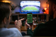 © DragonImages - Young adult men watching football match and checking betting app on smartphone, expressing male friendship and shared leisure. Useful for sports betting, gaming, entertainment marketing