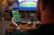 © DragonImages - Young adult man holding smartphone, checking sports betting app while watching match with friend, representing male friendship and fan engagement. For gaming, betting, sports marketing