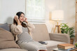 © bongkarn - Pregnant asian woman mother listening to music in headphones as sitting on sofa at home living room.