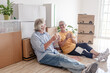 © luciano - An elderly couple moving into a new home, sitting on the floor for a break and a toast surrounded by moving boxes in an empty room. Concept of a new beginning, retirement, and relocation.