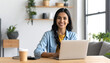 © Hermansyah - Smiling woman working on a laptop at a wooden desk in a bright home office setting, beverage and notebook nearby