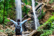 © torjrtrx - Rear view of a female traveler with a backpack raising her arms in joy and freedom while looking at a beautiful double waterfall. Female hiker celebrating success in a tropical rainforest in Thailand.
