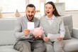 © Pixel-Shot - Happy young couple with refund form and piggy bank sitting on sofa at home