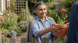 © Lambic - Old Woman giving poppy plant - Elderly woman smiling while handing flower pot over garden fence
