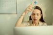 © DragonImages - Young adult Caucasian businesswoman cooling forehead with water bottle while working on laptop, showing heat stress and fatigue. Useful for workplace wellness, summer office safety
