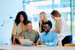 © we.bond.creations - Group of diverse business colleagues smiling and collaborating while looking at a laptop in a modern office