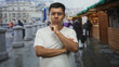 © Krakenimages.com - Young hispanic man in white tshirt holding his elbow and hand on chin while standing in a street market with wooden stalls and wet pavement; thoughtful reflection.