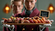 © Nick Freund - Zwei Kellner in roten Uniformen halten ein Tablett mit verschiedenen Sushi-Stücken und einem Kalorienhinweisschild.