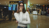Young woman wearing glasses biting her nails with hand to mouth gesture in an airport terminal near departure screens and queues; travel anxiety delay.