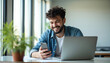 © Viktor - Happy man with curly hair works on laptop and mobile phone. Young guy smiles, uses tech devices for work or study. He sits at desk in modern office with big window.