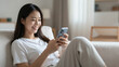 © CrazyJuke - Woman Relaxing with Smartphone: A young woman with a warm smile is engrossed in her smartphone while comfortably lounging on a sofa, embodying modern connection and leisure.