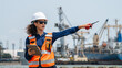 © VStudio - Woman wears a white hard hat and orange safety vest. She holds a tablet and points with a pen. She directs maritime freight operations at a commercial port. Cranes and ships appear in the background