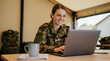 © Viktor - Smiling German soldier works on laptop inside tent camp. Female officer uses computer for operations. Pro woman in army uniform on duty with gear in background.