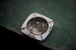 © Taufan - Close-up of a dirty metal ashtray filled with cigarette ash and residue, resting on a cracked green painted surface. Moody still life detail shot capturing everyday object texture and realism