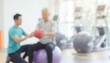 © al-sultan - A trainer assists a senior individual in a gym, focusing on strength training with a medicine ball while seated on a stability ball in a bright environment.