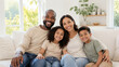 © Nadira - Joyful diverse family of four smiling warmly while relaxing together on a sofa in a bright, modern living room.