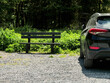 © mark_gusev - Black family car parked in a forest park parking lot next to a bench. Warm sunny day. Travel to nature concept. Fine day out for a group of people theme.