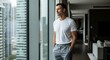 © Platon - A handsome man in a basic white t-shirt standing with his hands in his pockets inside a luxury penthouse apartment, looking thoughtfully out of a floor-to-ceiling window at the city.