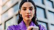 © v.stock - Portrait of a smiling young woman dress violet with a cheerful expression in a casual lifestyle setting holding smartphone modren building background.