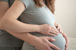 © Sk Elena - Pregnant woman with partner placing hands over her belly in shared intimate moment. Expecting mother and husband embracing from side in studio with neutral background. Mom-to-be celebrating shared