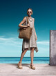 © Cumhur - Exquisite high fashion portrait of stunning Caucasian model wearing beige linen dress woven wedges and holding large fringed straw shoulder bag on wooden pier at exotic tropical beach resort