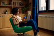 © Boris Jovanovic/Stocksy - Woman uses her laptop while seated in a green chair by the window