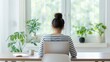 © Intelligent Horizons - Young woman s back view as she works on a laptop at a home desk bathed in natural daylight near plants and a window showcasing a calm and productive atmosphere