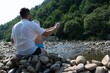 © Niko_Dali - A relaxing river view. Two people sit on rocks by a flowing river, enjoying the scenic landscape of lush green hills.