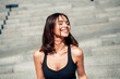 © deagreez - Smiling young woman in sports bra enjoying sunshine on outdoor steps after workout, healthy lifestyle and fitness concept
