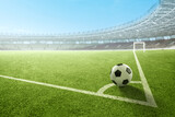 A black and white soccer ball on the corner kick line of a green field inside a large, empty stadium under a clear sky
