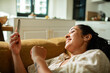 © Marko Geber - Smiling woman using smartphone on sofa at home