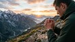 © Keo - A man kneels on a rocky mountain ledge, hands clasped in prayer, gazing at a breathtaking mountain vista during sunrise.
