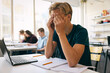 © Maskot - Tired teenage boy sitting with hands covered face at desk in classroom