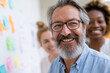 © lashkhidzetim - Happy man smiles at camera with glasses and beard. Behind him, colleagues smile, adding warmth to scene. Colorful sticky notes dot white wall, signaling teamwork. Ideal for business, collaboration