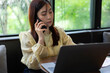 © Father_Studio - Asian businesswoman multitasking while working remotely in cafe, talking on smartphone and managing online business tasks.