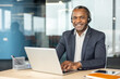 © Liubomir - Professional mature man working at a desk in an office, wearing a headset, smiling and looking at the camera while typing on a laptop, representing business communication and client service