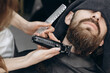 © MZaitsev - Professional female barber trimming beard of male client with electric clipper during grooming procedure in modern barbershop. Close up of hands and face.