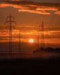 © Travel 'n' Lifestyle - View of high voltage power lines and electricity pylons silhouetted against a vibrant orange sunset sky with clouds over a rural field in Kyjov, South Moravian Region, Czechia.