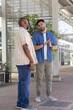 © wavebreak3 - Two African American males talking, gesturing under covered plaza, holding phone and wearing watch