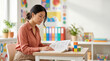 © Vera D - Focused young asian female teacher reading open textbook at desk in bright modern elementary classroom interior, woman mentor studying book in primary school background