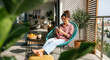 © pradeep - Smiling Young Woman Relaxing Barefoot with Smartphone in Comfortable Chair on Sunlit Modern Balcony with City View