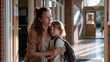 © Tania - Kindergarten teacher kneeling beside anxious five year old clinging to parent's leg at classroom entrance on first day, backpack nearly as tall as child, morning sunlight crossing doorway threshold