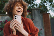 © SHOTPRIME STUDIO - Joyful woman outdoors holds a phone, smiling with curly hair in a red sweater. Bright natural light and greenery frame a candid lifestyle moment focused on happiness and everyday joy